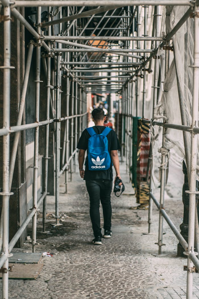 Man walking under scaffolding in city, carrying a helmet and wearing a backpack.